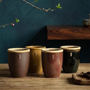 Puede incluir: Cuatro tazas de cerámica en tonos marrón, amarillo y negro dispuestas sobre una superficie de madera. Cada taza tiene un borde de color claro y un pequeño diseño en relieve. Las tazas son cilíndricas y parecen hechas a mano.