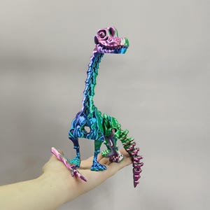 Puede incluir: Figura de esqueleto de dinosaurio articulada en tonos azules, verdes y morados. El juguete se sostiene en una mano, mostrando su diseño detallado y acabado iridiscente. El dinosaurio tiene un cuello y una cola largos.