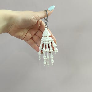 Può includere: Un portachiavi a forma di mano scheletrica bianca con un anello argentato. La mano in miniatura è una replica dettagliata di una mano umana, con singole ossa visibili. Il portachiavi è un accessorio unico, perfetto per chi è interessato all'anatomia o all'estetica gotica.