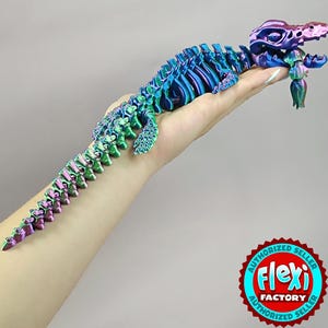 Op de afbeelding: Een veelkleurig, beweeglijk dinosaurus speelgoed in tinten blauw, groen en paars. Het speelgoed wordt in een hand gehouden, waarbij het flexibele ontwerp wordt getoond. Het Flexi Factory-logo is rechtsonder zichtbaar.