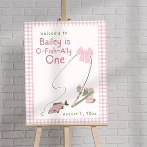 May include: A welcome sign on an easel with a pink gingham border. The sign reads "Welcome to Bailey is O-Fish-Ally One" with a fishing rod illustration and the date August 11, 20xx. The sign is on a wooden easel in front of a white brick wall.