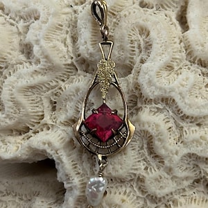 May include: A gold-toned pendant with a square, red gemstone at its center, framed by an openwork design. A small, irregular pearl dangles below. The pendant is set against a textured, off-white background.