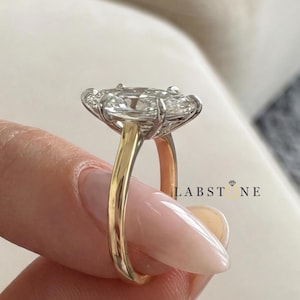 May include: A gold and silver engagement ring with a large, oval-shaped clear stone. The ring has a gold band and a silver setting that holds the stone. The ring is held up by a person's fingers.