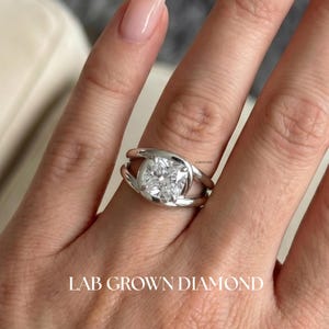 Puede incluir: Primer plano de un anillo de plata con un diamante cuadrado cultivado en laboratorio. El anillo tiene un diseño único con una caña dividida que se curva alrededor del diamante. El texto "LAB GROWN DIAMOND" está en la parte inferior de la imagen.