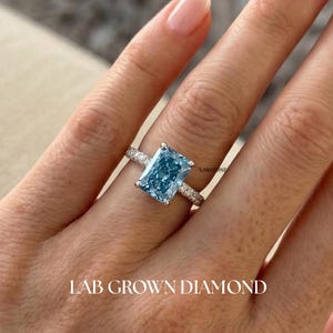 Puede incluir: Un anillo de plata con un gran diamante azul claro rectangular cultivado en laboratorio. La banda está adornada con piedras más pequeñas y transparentes. Las palabras "LAB GROWN DIAMOND" están impresas en la parte inferior de la imagen. El anillo está en un dedo.