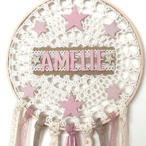 Könnte beinhalten: Handgefertigter Traumfänger mit Holzreifen und weißem Häkelnetz. Rosa Sterne und der Name "AMELIE" sind zu sehen. Verziert mit rosa und cremefarbenen Bändern und Spitze, ein bezauberndes Dekorationsobjekt für Kinderzimmer oder Schlafzimmer.