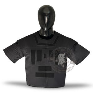 May include: Black tactical shirt with Velcro panels and short sleeves, displayed on a mannequin. The shirt has a crew neck and is designed for protection. A logo is visible on the right side of the shirt.