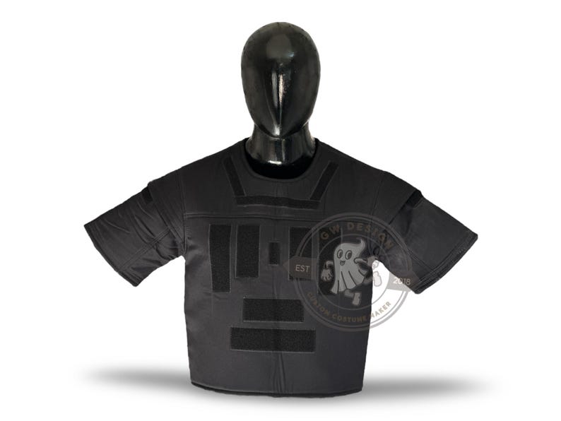 May include: Black tactical shirt with Velcro panels and short sleeves, displayed on a mannequin. The shirt has a crew neck and is designed for protection. A logo is visible on the right side of the shirt.