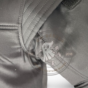 May include: Close-up of a grey garment with visible stitching and a textured fabric. The image includes a portion of a sleeve and shoulder area. A logo with a ghost character and the text "EST 2018" is visible.