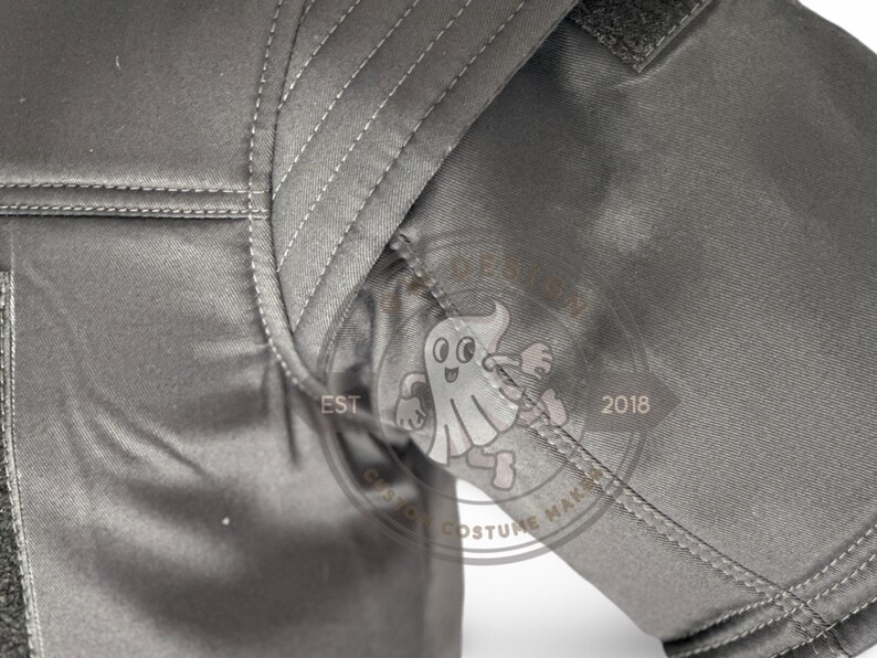 May include: Close-up of a grey garment with visible stitching and a textured fabric. The image includes a portion of a sleeve and shoulder area. A logo with a ghost character and the text "EST 2018" is visible.