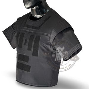 May include: Black tactical vest with short sleeves, featuring multiple rectangular velcro patches on the front. The vest is displayed on a mannequin. The image includes a logo with the text "CUSTOM COSTUME MAKER EST 2018".