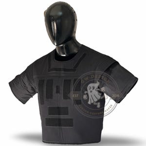 May include: Black tactical vest on a mannequin. The vest has several rectangular panels and shoulder pads. It features hook-and-loop fastener areas. The mannequin is black, and the background is white.
