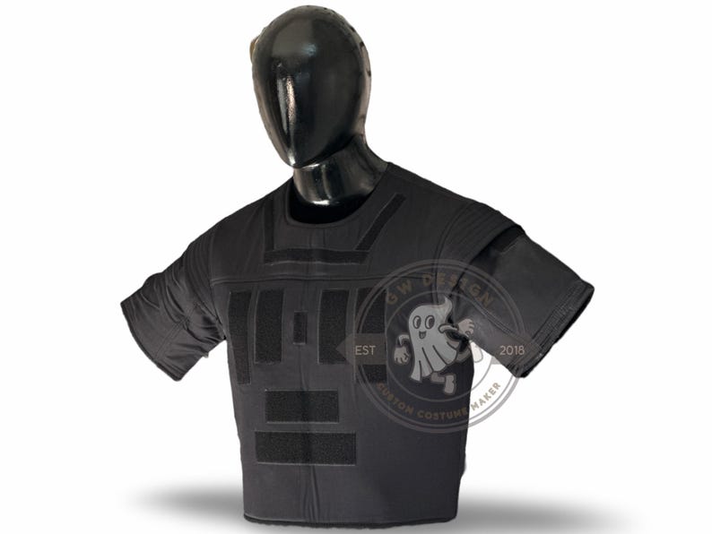 May include: Black tactical vest on a mannequin. The vest has several rectangular panels and shoulder pads. It features hook-and-loop fastener areas. The mannequin is black, and the background is white.