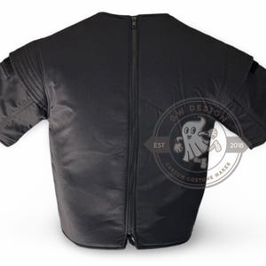 May include: Black short-sleeved garment with a full-length zip at the back. The garment features padded shoulders and a small pocket on each sleeve. A circular logo with a ghost graphic is visible.