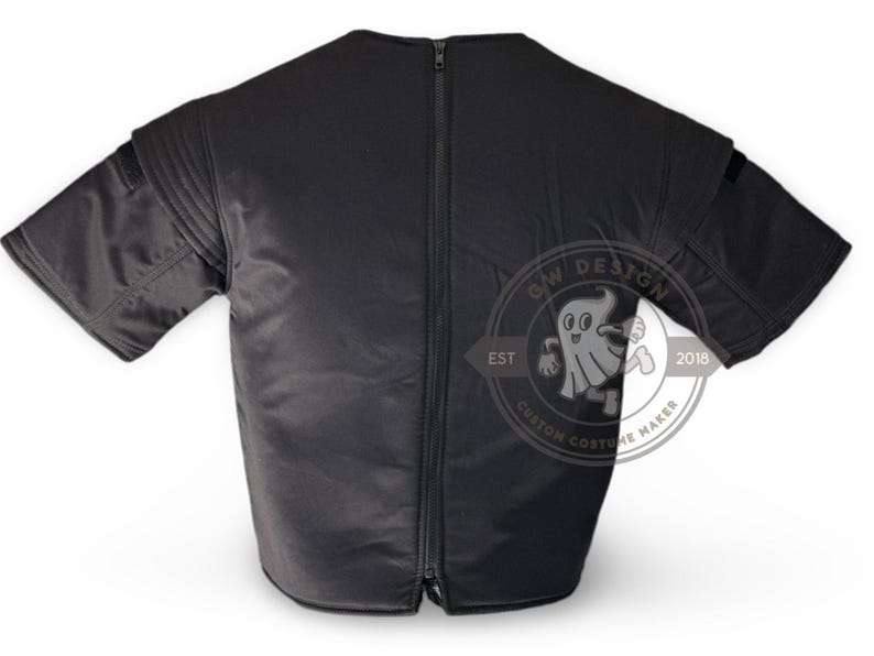 May include: Black short-sleeved garment with a full-length zip at the back. The garment features padded shoulders and a small pocket on each sleeve. A circular logo with a ghost graphic is visible.