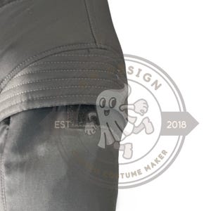 May include: A close-up of a section of a dark grey garment, possibly a jacket or coat, with quilted stitching details. The image includes a portion of a logo with the text "EST" and a cartoon ghost figure.