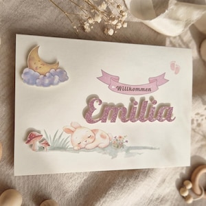 Könnte beinhalten: Eine rechteckige weiße Karte mit dem Namen Emilia in glitzernder rosa Schrift. Das Design zeigt einen schlafenden Hasen, eine Mondsichel auf einer Wolke, Pilze und ein rosa Banner mit dem Text Willkommen. Kleine Babyfußabdrücke sind zu sehen.