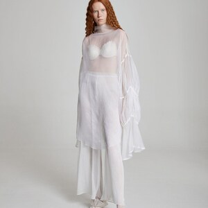 May include: A sheer white ensemble featuring a high-neck top with voluminous sleeves, paired with matching pants and a skirt overlay. A white bra is visible beneath the top. The outfit is completed with white shoes. The overall style is ethereal and flowing.