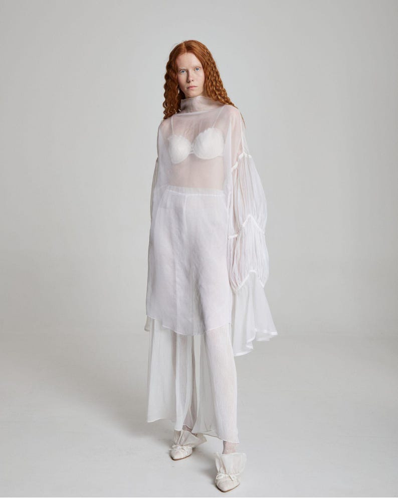 May include: A sheer white ensemble featuring a high-neck top with voluminous sleeves, paired with matching pants and a skirt overlay. A white bra is visible beneath the top. The outfit is completed with white shoes. The overall style is ethereal and flowing.