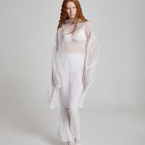 May include: A sheer white ensemble featuring a high-neck top with bell sleeves, paired with matching pants. The outfit includes a white bra and is completed with white shoes. The model has long, wavy red hair.