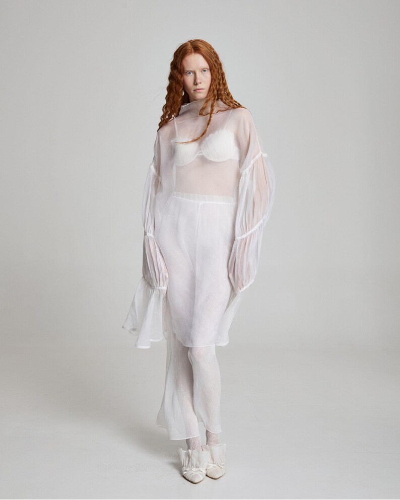 May include: A sheer white ensemble featuring a high-neck top with bell sleeves, paired with matching pants. The outfit includes a white bra and is completed with white shoes. The model has long, wavy red hair.