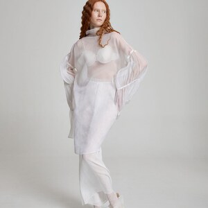 May include: A sheer white top with long, flared sleeves and a high neckline is layered over a white bra with a unique, textured design. The outfit includes a matching white skirt and pants, paired with white shoes. The overall look is ethereal and avant-garde.