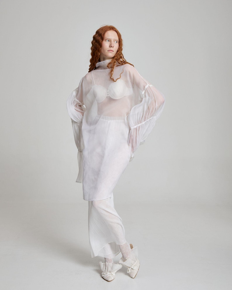 May include: A sheer white top with long, flared sleeves and a high neckline is layered over a white bra with a unique, textured design. The outfit includes a matching white skirt and pants, paired with white shoes. The overall look is ethereal and avant-garde.