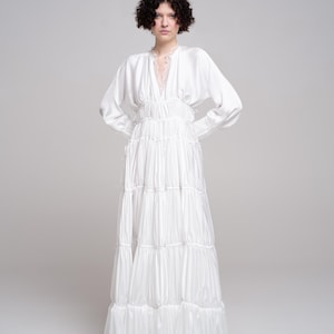 May include: A floor-length white satin dress featuring long sleeves, a deep V-neckline with delicate lace trim, and a gathered bodice. The skirt is constructed with multiple horizontal tiers, creating a voluminous silhouette with a soft sheen.