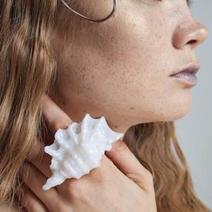 Pode incluir: Um anel de concha branco. O anel é um design esculpido e texturizado que se assemelha a uma concha. O anel é usado em um dedo. O fundo é de cor neutra.