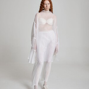 May include: A sheer white ensemble featuring a high-neck top with flared sleeves, a matching skirt, and leggings. The outfit includes a white bra and is paired with white shoes. The overall look is ethereal and flowing.