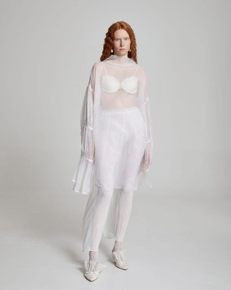 May include: A sheer white ensemble featuring a high-neck top with flared sleeves, a matching skirt, and leggings. The outfit includes a white bra and is paired with white shoes. The overall look is ethereal and flowing.