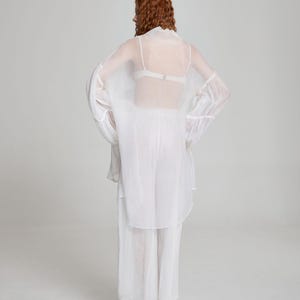 May include: A sheer white outfit consisting of a long-sleeved top, pants, and a matching overcoat. The outfit is worn over a white bra. The model has red hair. The outfit is made of a lightweight, flowing fabric.