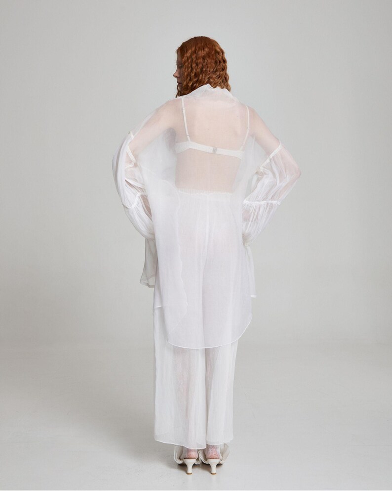 May include: A sheer white outfit consisting of a long-sleeved top, pants, and a matching overcoat. The outfit is worn over a white bra. The model has red hair. The outfit is made of a lightweight, flowing fabric.