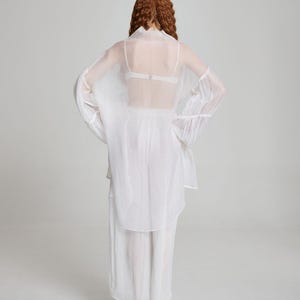 May include: A sheer white outfit consisting of a long-sleeved top with flared sleeves, a matching skirt, and a delicate bra. The outfit is made of a lightweight, flowing fabric, creating a soft, ethereal look. The model is facing away from the camera.
