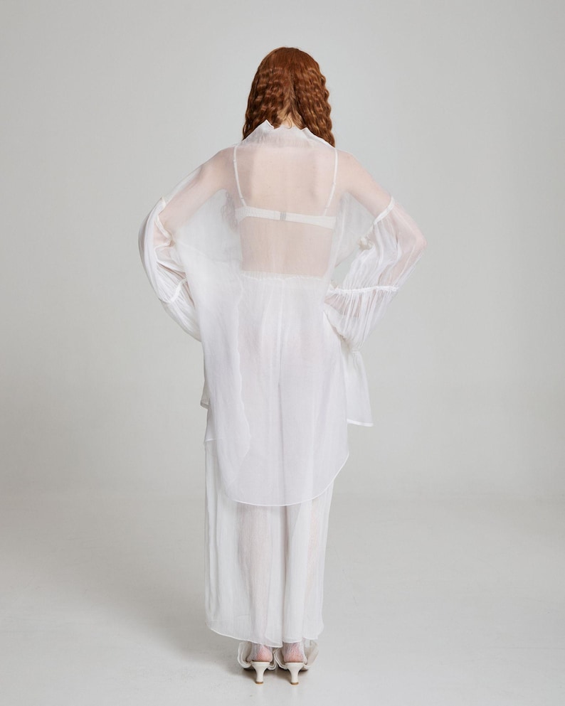 May include: A sheer white outfit consisting of a long-sleeved top with flared sleeves, a matching skirt, and a delicate bra. The outfit is made of a lightweight, flowing fabric, creating a soft, ethereal look. The model is facing away from the camera.