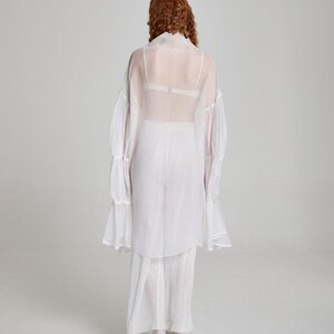 May include: A white, sheer, long-sleeved blouse with ruffled cuffs and a matching skirt. The outfit is worn over a white bra. The model is facing away from the camera, with red hair.