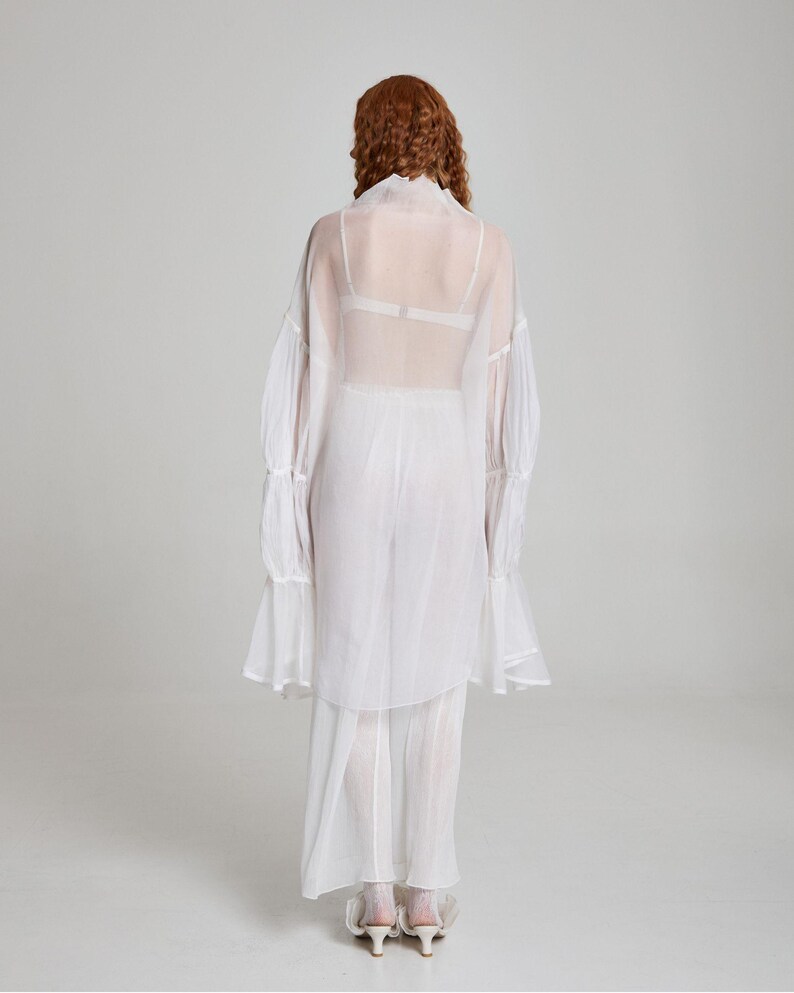May include: A white, sheer, long-sleeved blouse with ruffled cuffs and a matching skirt. The outfit is worn over a white bra. The model is facing away from the camera, with red hair.