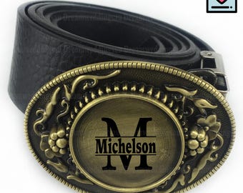 May include: A black leather belt with a decorative, oval-shaped buckle. The buckle is bronze-coloured with floral and leaf designs around the edges. The centre of the buckle has the letter "M" and the name "Michelson" engraved.