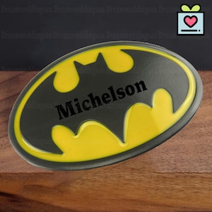 Peut inclure: Un logo Batman noir et jaune ovale avec le nom "Michelson" en noir. Le logo a un contour jaune et un intérieur noir. L'arrière-plan est une surface en bois.