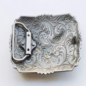 May include: Silver-toned rectangular belt buckle with intricate floral and scrollwork engraving. The buckle features a classic western design with a sturdy metal loop and pin for secure fastening. The edges have a decorative scalloped design.