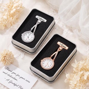 May include: Two nurse fob watches in rectangular metal gift boxes with black foam inserts. One watch is silver-toned with the name Alison engraved on the pin, and the other is rose gold-toned with the name Rachel engraved on the pin.