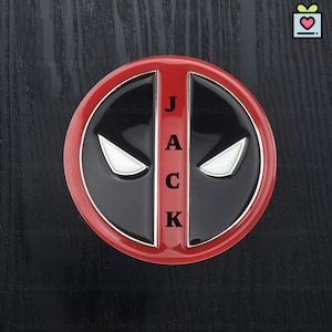 May include: A circular emblem in red, black, and white, displaying the Deadpool mask design. The word "JACK" is vertically aligned in white lettering. The emblem has a shiny finish and is set against a dark wood grain background.