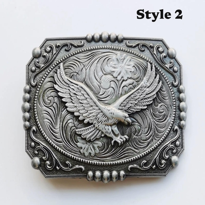 May include: Silver-toned metal belt buckle with an embossed eagle design, wings outstretched. The buckle has an ornate, floral-inspired border and the text "Style 2" in the upper right corner. The eagle is the focal point.