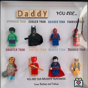 Puede incluir: Una obra de arte enmarcada con figuras de superhéroes de Lego, incluyendo a Hulk, Batman y Spider-Man. El texto dice "Papá, tú eres..." con comparaciones temáticas de superhéroes. El texto inferior dice "Eres nuestro superhéroe favorito".