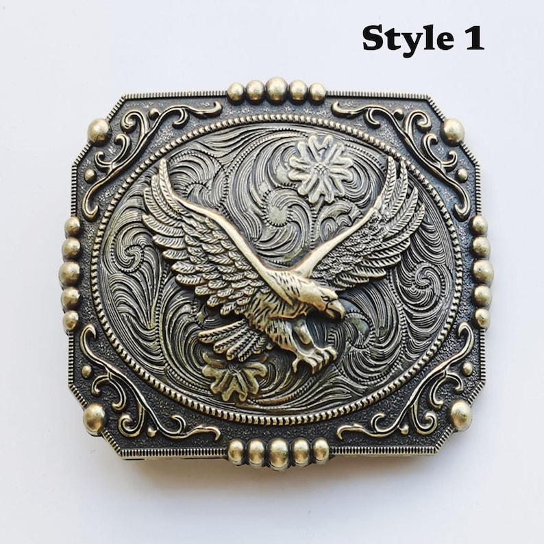 May include: A rectangular, antique-style belt buckle with an embossed eagle design. The eagle is centered, with outstretched wings, against a detailed floral and scrollwork background. The buckle is metallic with a bronze finish, and the text "Style 1" is in the upper right corner.