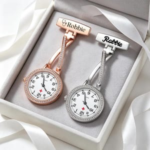 Può includere: Due orologi da infermiere, uno in oro rosa e uno in argento, con quadrante bianco, numeri neri, un simbolo di croce rossa e una lunetta con cristalli. Ogni orologio ha una spilla incisa con il nome Robbie e un'icona a forma di stetoscopio.