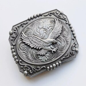 May include: Silver-toned metal belt buckle with an eagle design. The buckle features an embossed eagle with outstretched wings, surrounded by ornate scrollwork and a beaded border. The rectangular buckle has a decorative, detailed design.