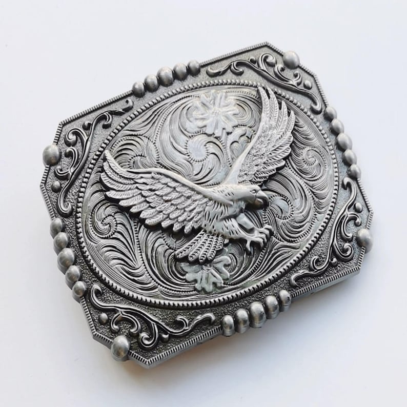 May include: Silver-toned metal belt buckle with an eagle design. The buckle features an embossed eagle with outstretched wings, surrounded by ornate scrollwork and a beaded border. The rectangular buckle has a decorative, detailed design.