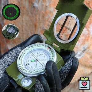 May include: A green military-style compass with a white dial, black markings, and a clear sighting lens. The compass has a built-in bubble level and a rotating bezel. The image shows the compass open, revealing the internal components. The compass is held by a person wearing black gloves.