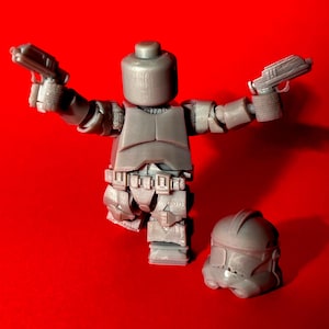 Könnte beinhalten: Eine graue Plastik-Actionfigur eines Star Wars Klonkriegers, der zwei Blaster hält. Die Figur trägt einen Helm und eine Rüstung. Ein separater grauer Plastik-Klonkriegerhelm ist ebenfalls enthalten.